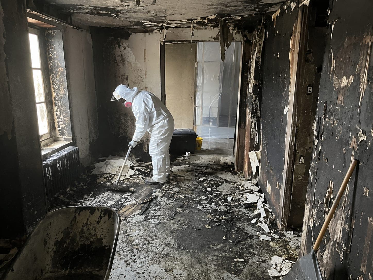 Fachkraft in Schutzanzug und Atemschutz räumt einen rußgeschwärzten Innenraum nach einem Brand aus